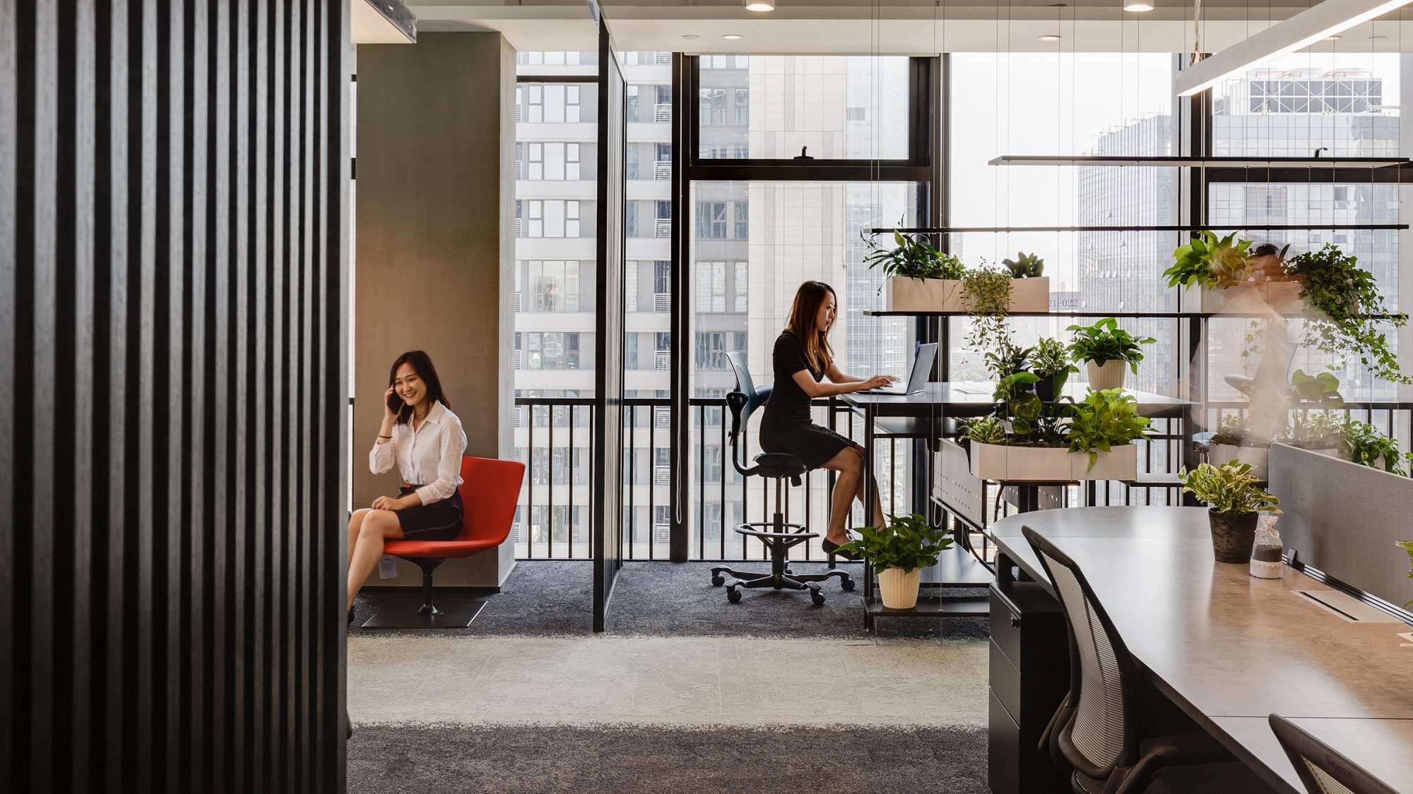 woman working at desk surrounded by plants and woman sitting in chair on phone