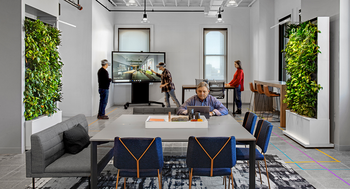 man working at desk, man presenting using screen and woman standing small