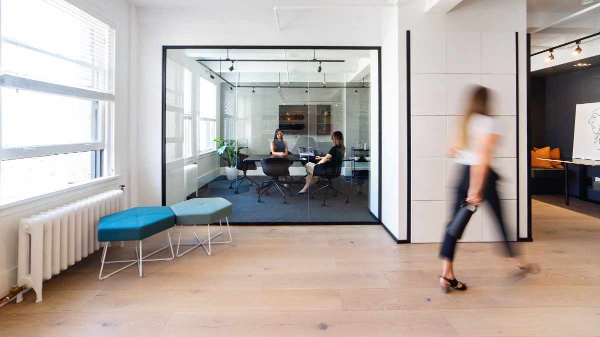 people collaborating in meeting room and woman walking through workplace