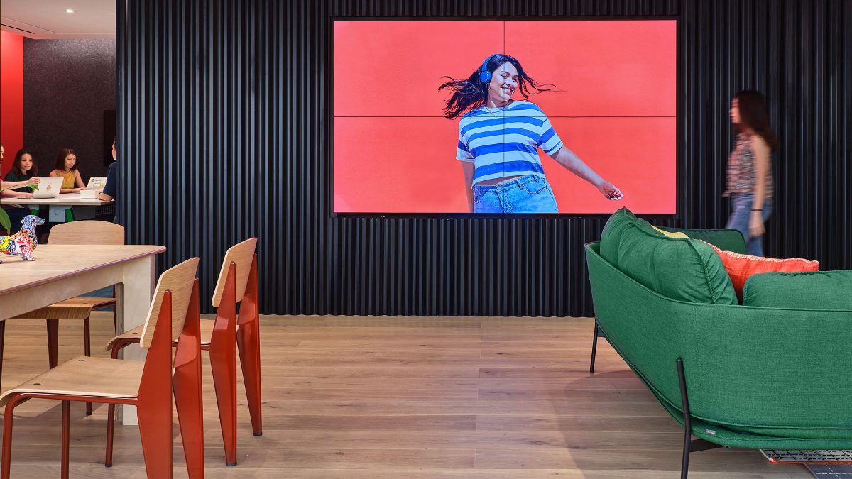 woman walking in front of av wall and people seen to be collaborating at back