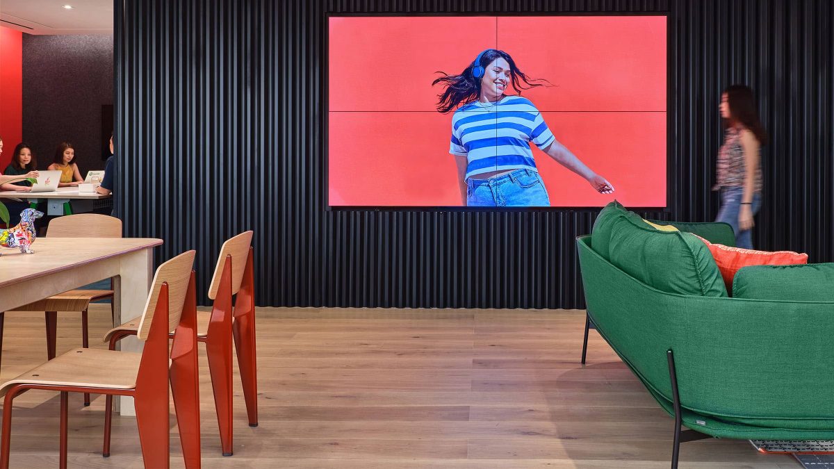 woman walking in front of av wall and people seen to be collaborating at back